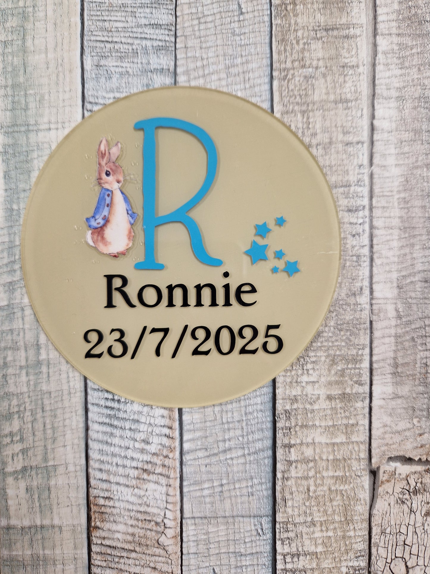 Round beige plaque with a blue letter 'R', name 'Ronnie', date '23/7/2025', and a rabbit illustration on a wooden background.