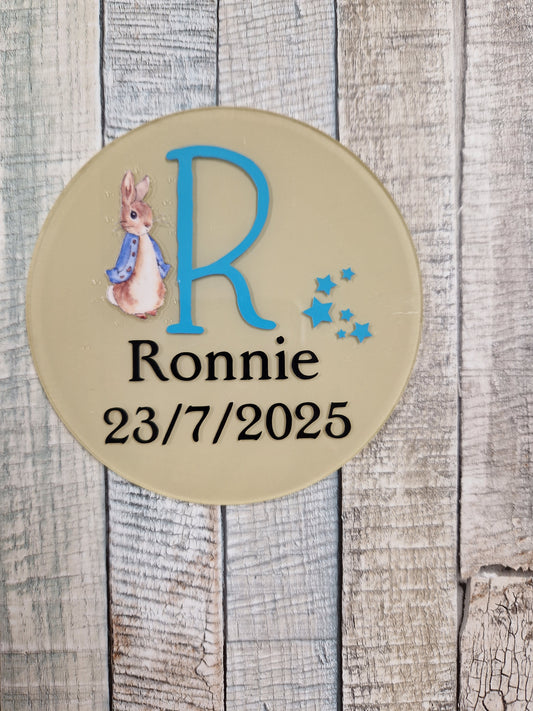 Round beige plaque with a blue letter 'R', name 'Ronnie', date '23/7/2025', and a rabbit illustration on a wooden background.